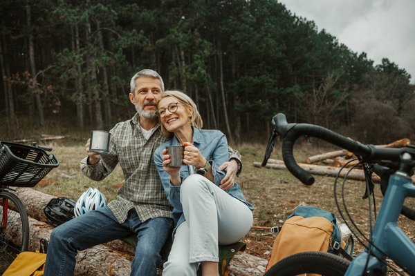 Two people with bicycles take a break in a woodland setting, sitting on a fallen tree and holding mugs.
